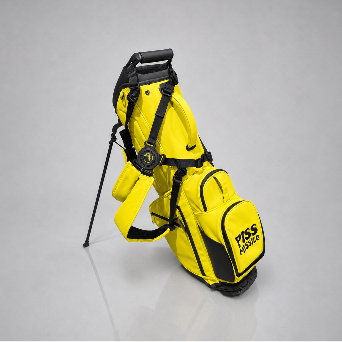 A bright yellow and black stand golf bag with a "PISS MISSILE" graphic on the front pocket, standing on a reflective gray studio floor.
